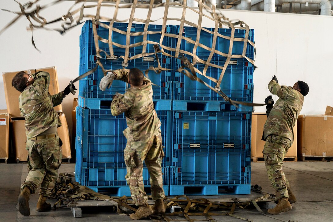 Airmen and soldiers throw a net onto a cargo pallet with cardboard boxes in the background.