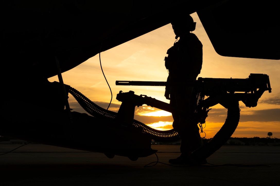 An airman stands next to a weapon attached to an aircraft on a tarmac under a sunlit sky.