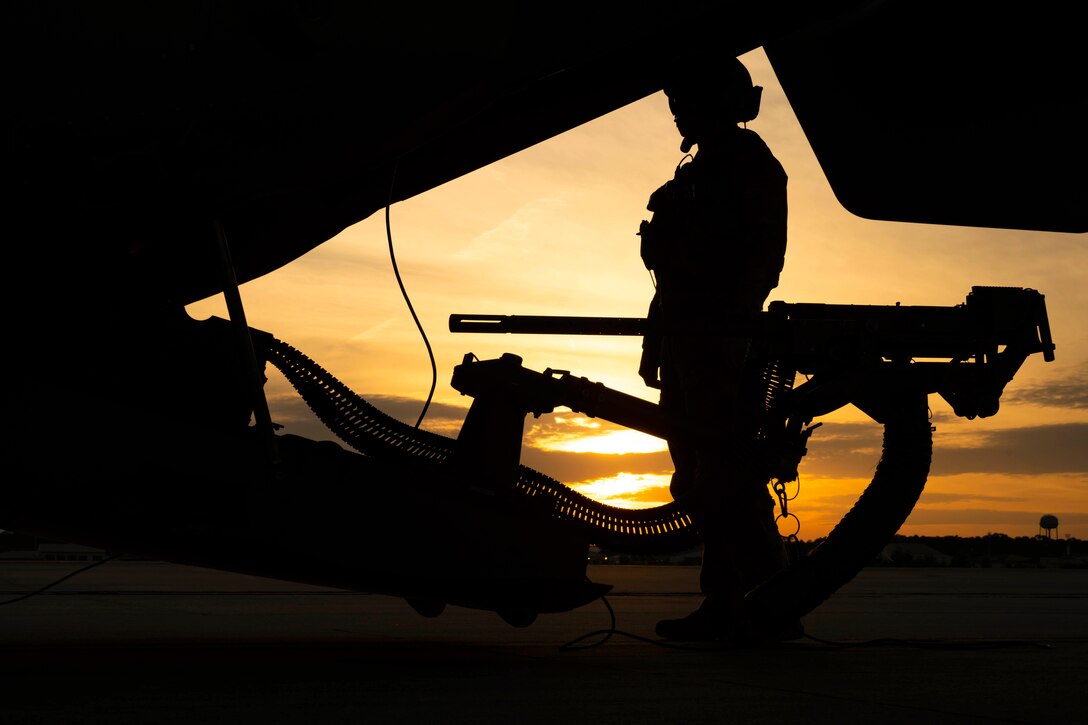 An airman stands next to a weapon attached to an aircraft on a tarmac under a sunlit sky.