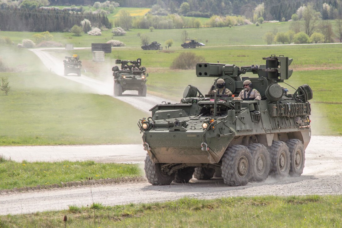 Soldiers drive armor vehicles on dirt roads surrounded by greenery.
