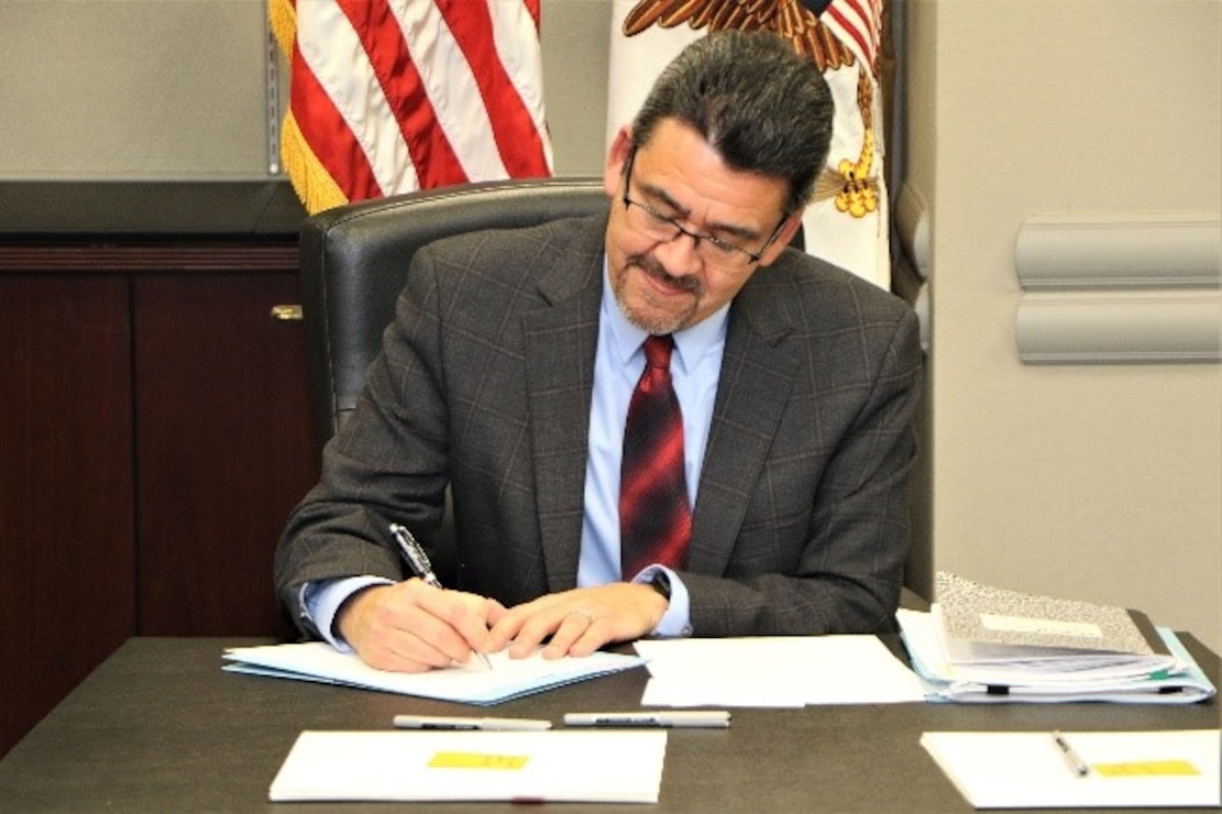 A man in a business suit signs documents at the head of a conference room table.
