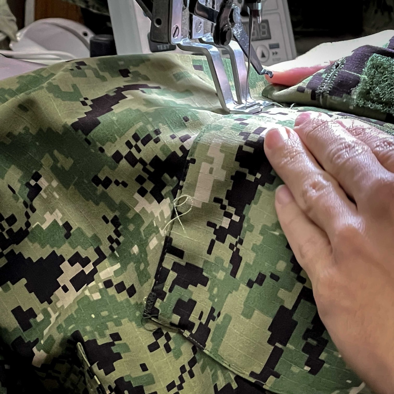 A close-up photo shows the hands of a person using a sewing machine to stitch a military uniform.