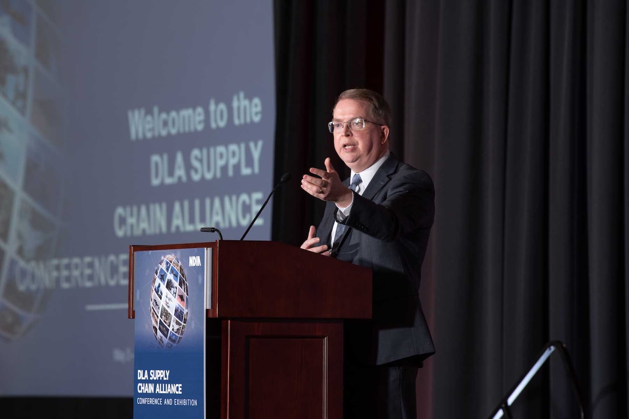 A man wearing business attire speaks while standing behind a lectern with a presentation in the background.