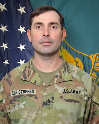 Man in U.S. Army uniform standing in front of two flags.