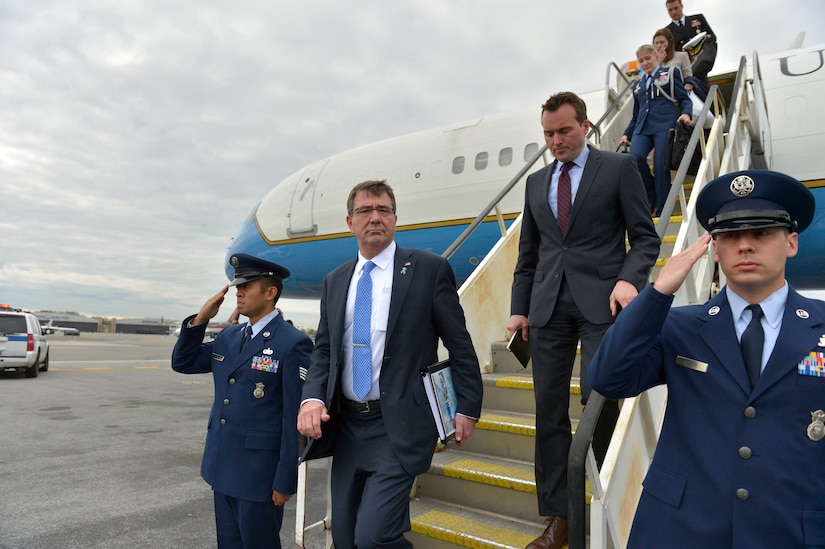 A man in a suit disembarks an airplane as men in uniform salute.