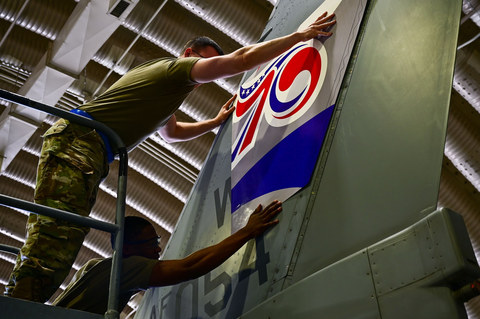 Staff Sgt. Matthew Campbell, 8th Maintenance Squadron aircraft structural maintenance craftsman (top), and Senior Airman Connor Young, 8th MXS aircraft structural maintenance, journeyman apply a 70th anniversary heritage tail flash decal at Osan Air Base, Republic of Korea, May 1, 2023. The decal comes in eight pieces, four for each side of the aircraft, and must be measured, leveled and applied securely on the aircraft. (U.S. Air Force photo by Tech. Sgt. Timothy Dischinat)