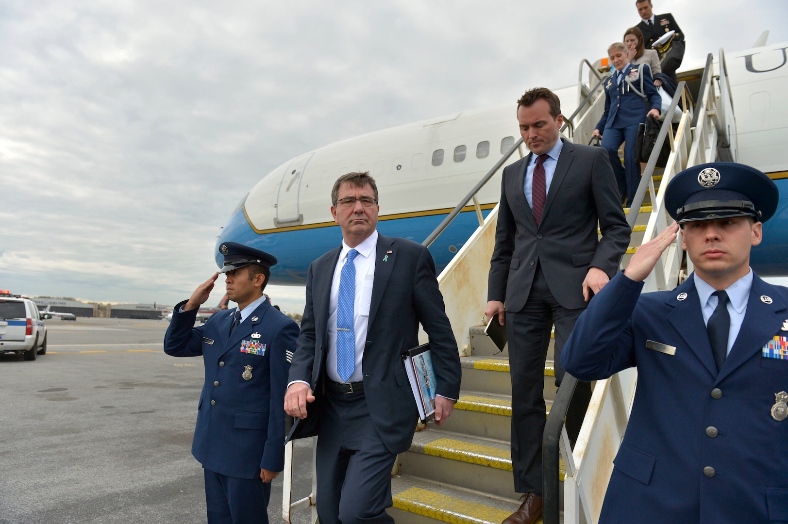 A man in a suit disembarks an airplane as men in uniform salute.