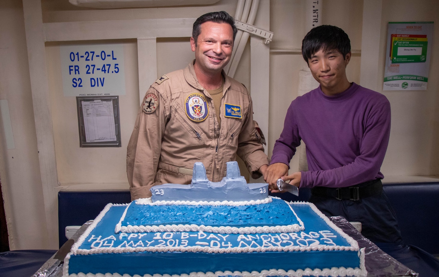 Capt. Daniel Keeler, commanding officer of amphibious transport dock USS Anchorage (LPD 23), left, and Aviation Boatswain’s Mate (Fuel) Airman Joel Kim, from Anchorage, Alaska, participate in a cake-cutting ceremony for the 10th anniversary of Anchorage’s commissioning, May 7, 2023.