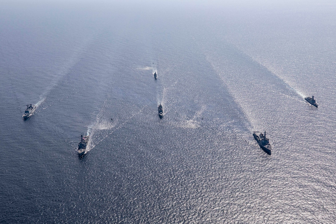 Helicopters fly above a group of ships sailing in formation.