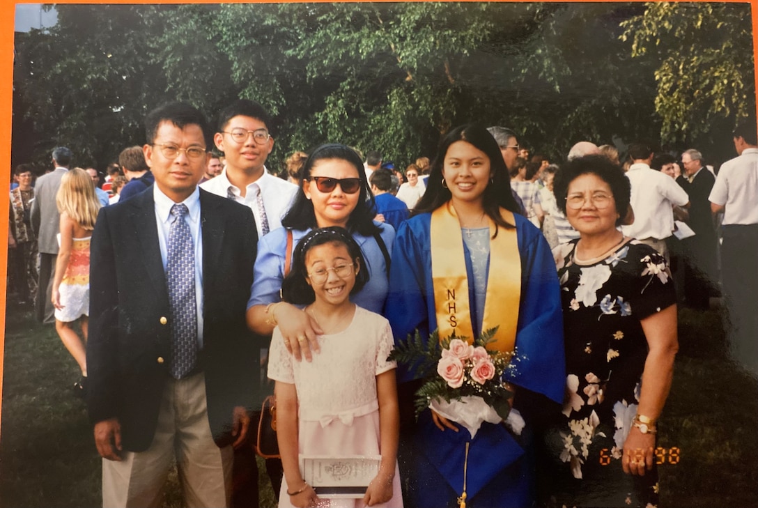 A family consisting of two men and four women, poses together outside with smiles on their faces. A young women in a blue and gold graduation gown and holding pink flowers is the center of attention; as she is being celebrated for her high school graduation.