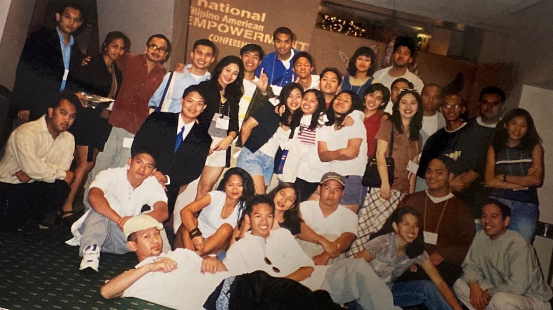 A large group of teenagers posed together in a large group. Some are hugging and smiling in front of a brown wall with a conference logo embroidered on it.