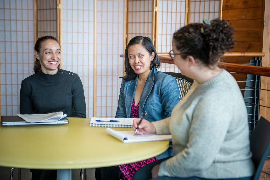 Three women sit at circular yellow table with white note pads a pen in from of them. Behind them is a brown wood and white wall.