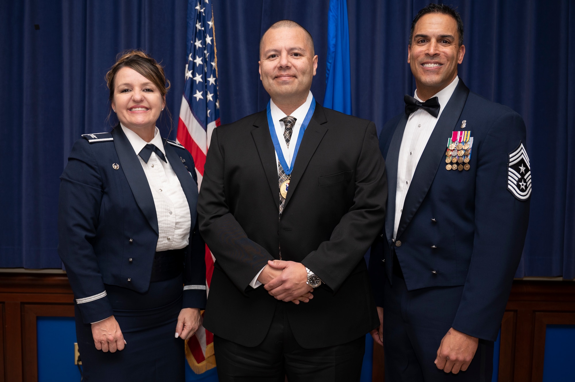 Three people pose for a photo in dress blues and business attire. the Center member is wearing an honorary medal.