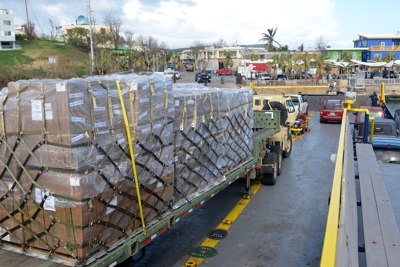 A military truck carries several pallets containing food and water to people.