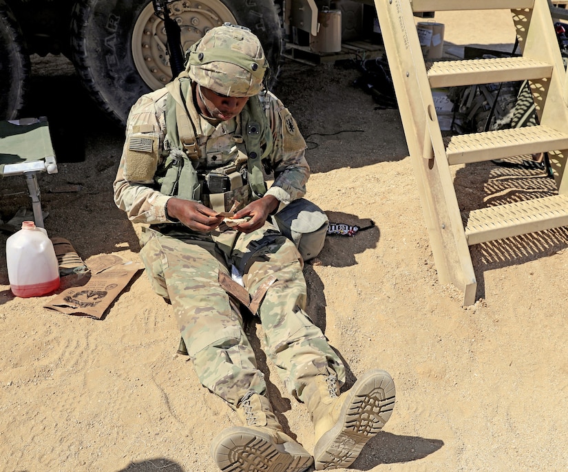 A uniformed service member eats a prepackaged meal while sitting in the sand.