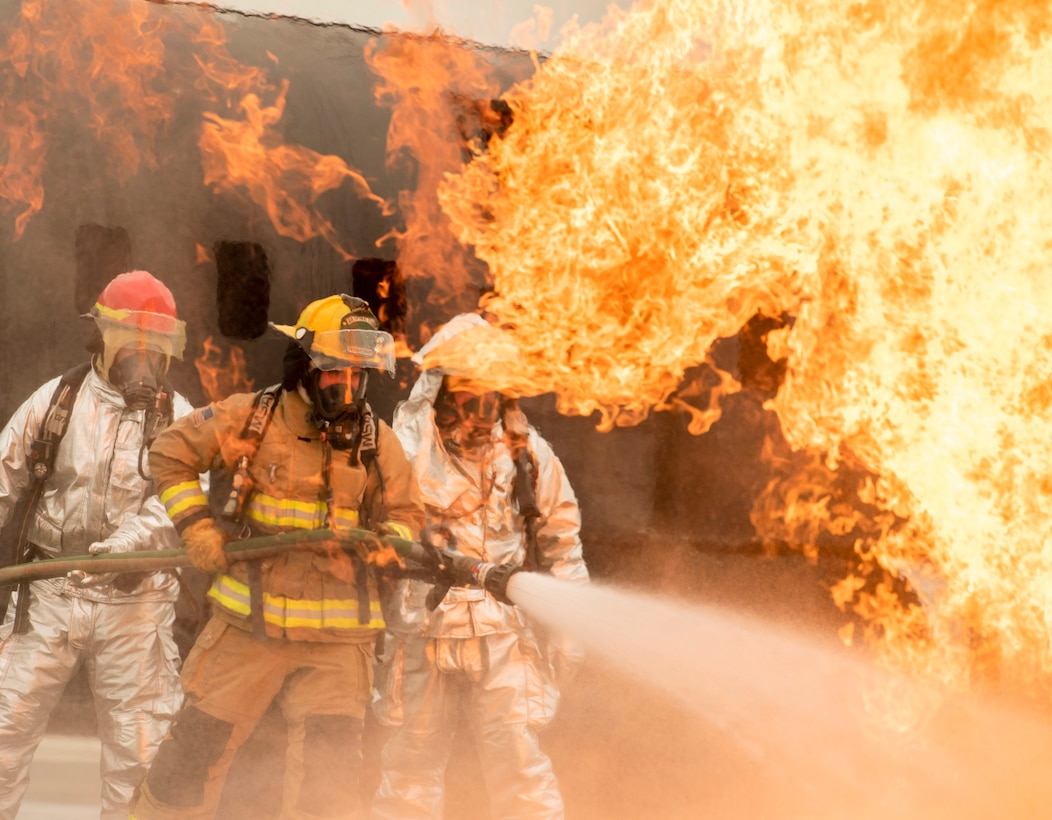 Three firefighters in protective gear use a water hose to put on a fire as a burning aircraft sits in the background.