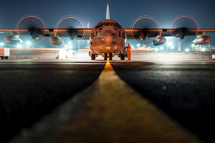 A service member stands next to an aircraft parked on a tarmac in the dark illuminated by streetlights.