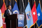 A man speaks at a podium. A row of military flags is behind him.