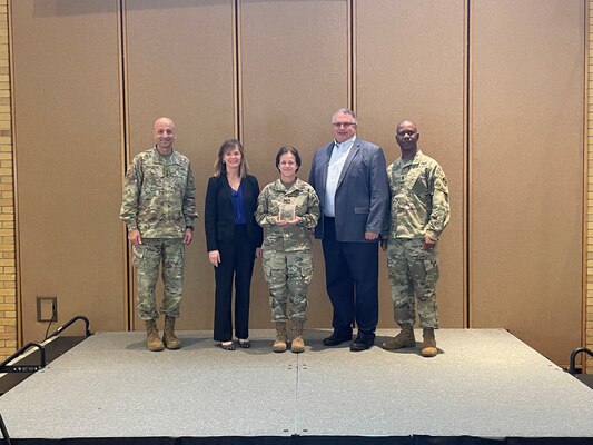 Four people, two women and three men, pose in front of a brown wall in a fluorescent lit room while receiving an award.