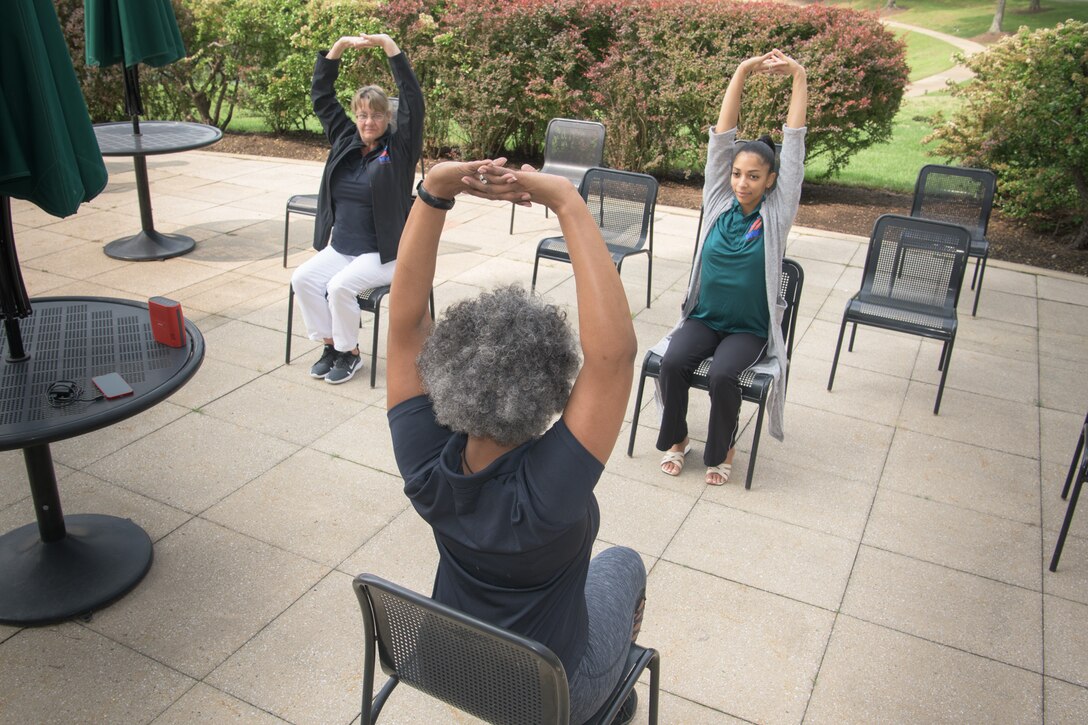 Three women sit in chairs in a yoga pose outside on a patio.