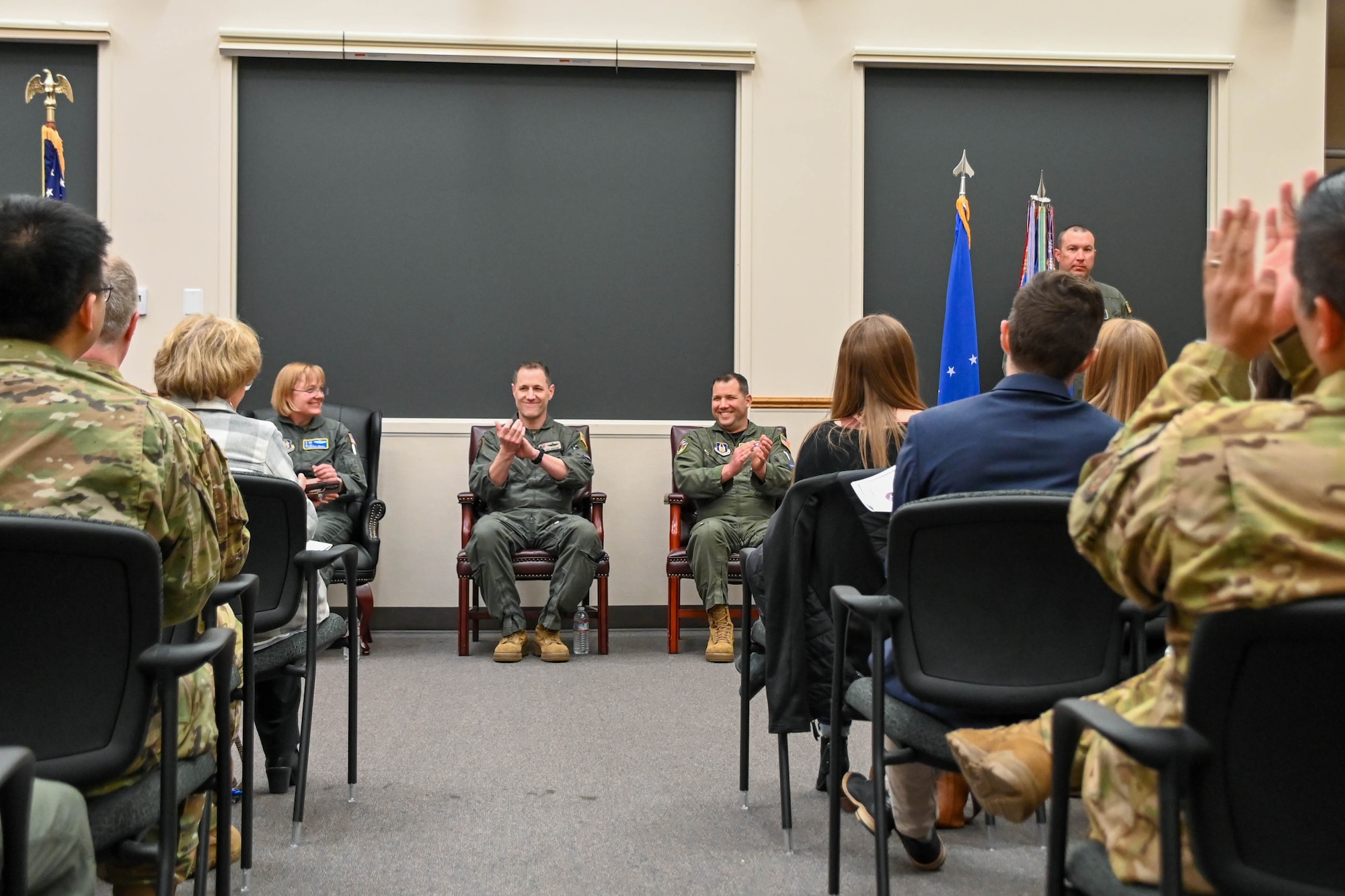 A civilian and military audience applaud the speaker as he stands at a podium at the front of the room and three people in flight suits sit in chairs at the front of the room also clap.