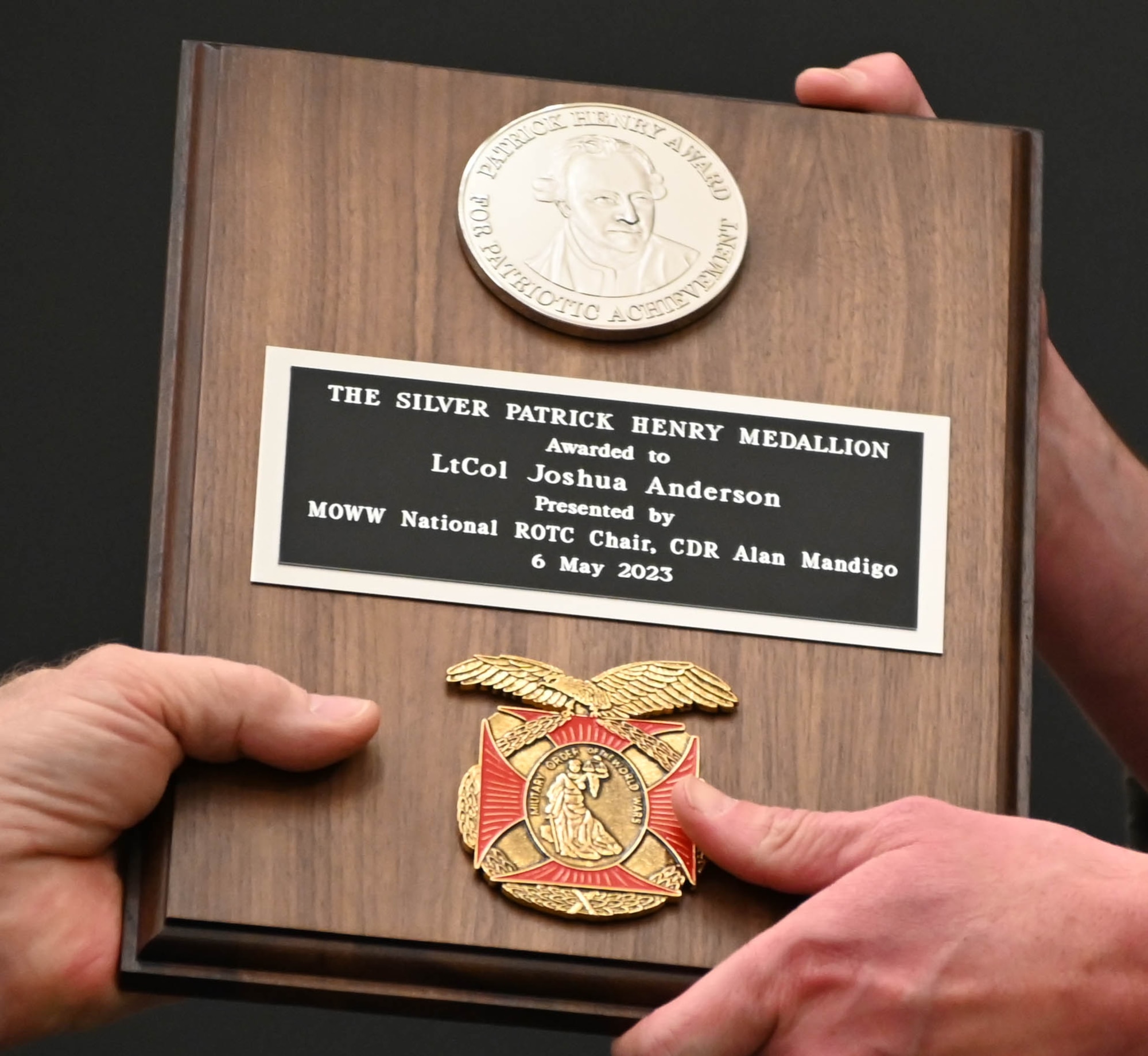 A pair of hands holding a wooden plaque that reads "The Silver Patrick Henry Medallion: Awarded to LtCol Joshua Anderson as presented by MOWW National ROTC Chair, CDR Alan Mandigo dated 6 May 2023"