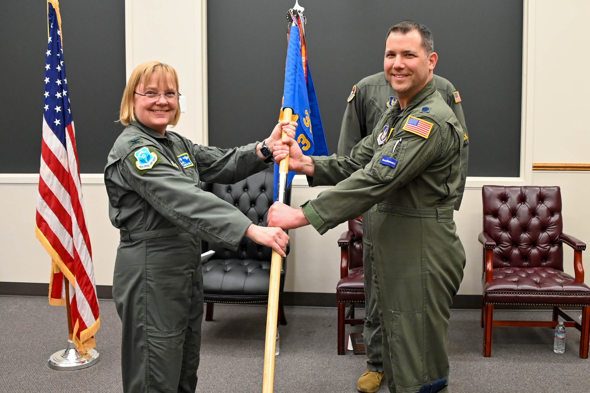 A woman in a green flight suit hands the blue flag to a man in a green flight suit in front of an American Flag.