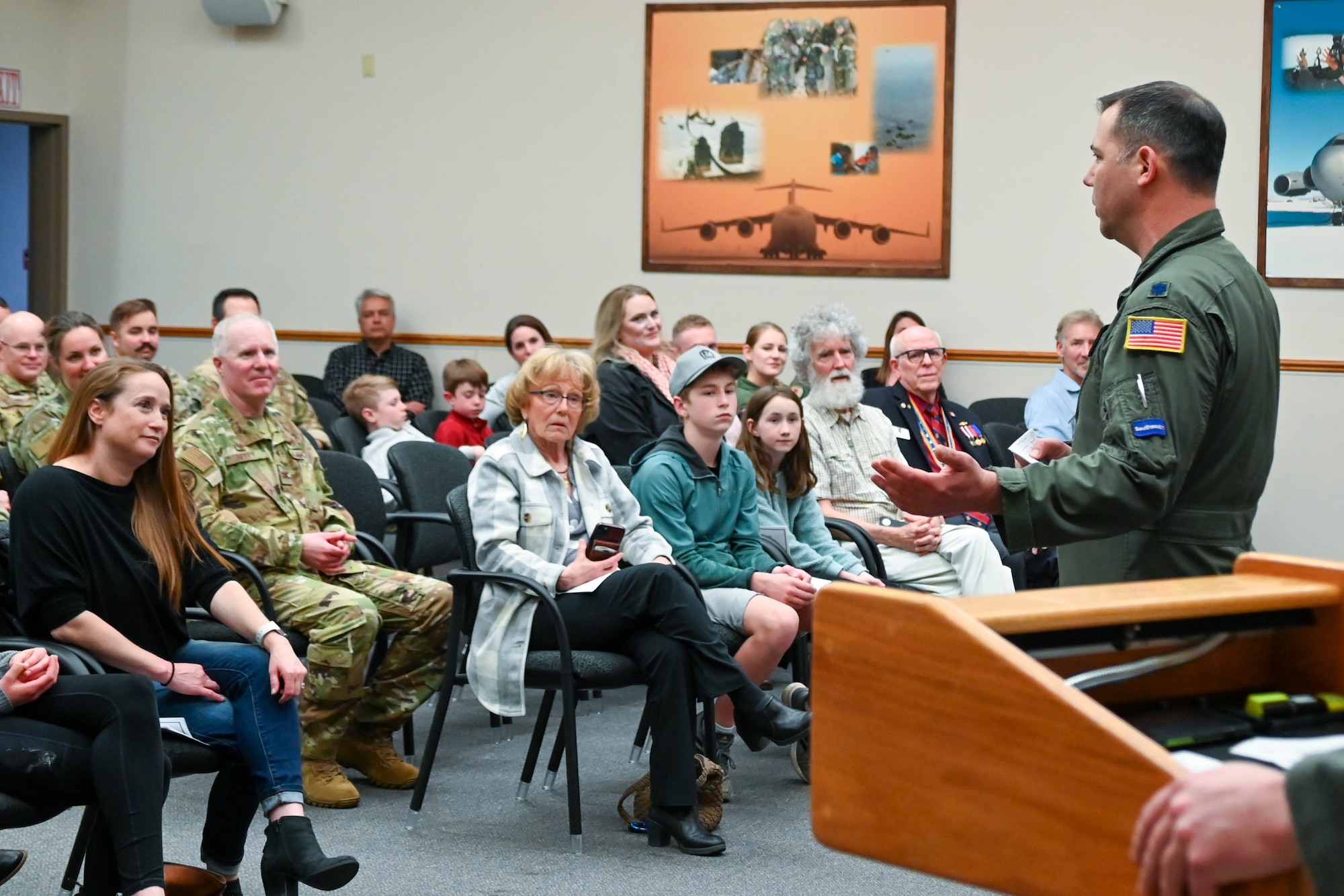 A man wearing a green flight suit gives a speech to a mixed civilian and military audience.