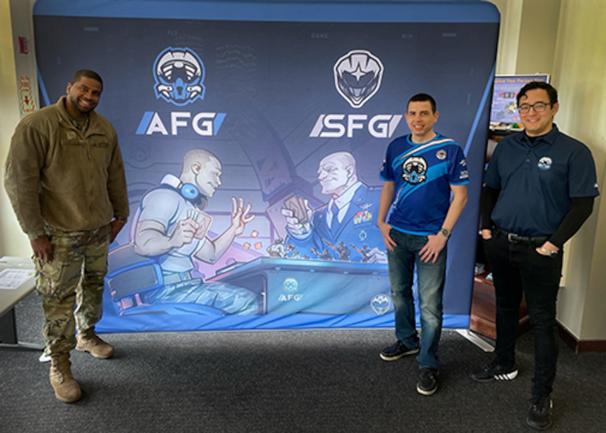 three men stand in front of a giant Air Force gaming banner. the banner features original artwork of an enlisted member playing a board game with a general officer.