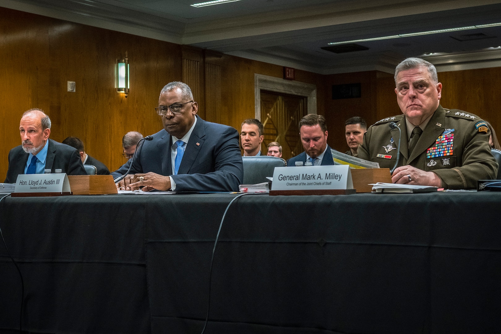 Three men, one in military uniform, sit at a long table. Other people are in the background.