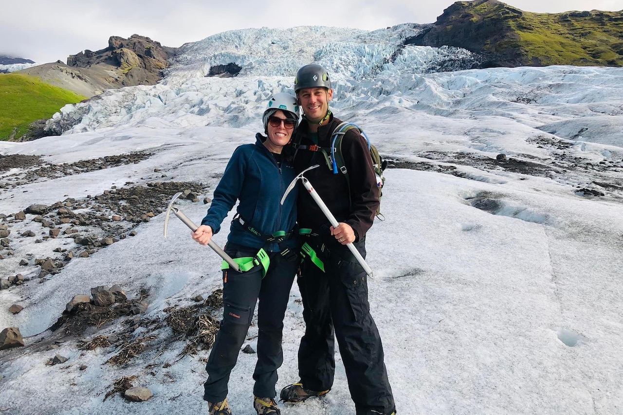 A couple pose on a snow-covered mountain area with ice climbing tools.