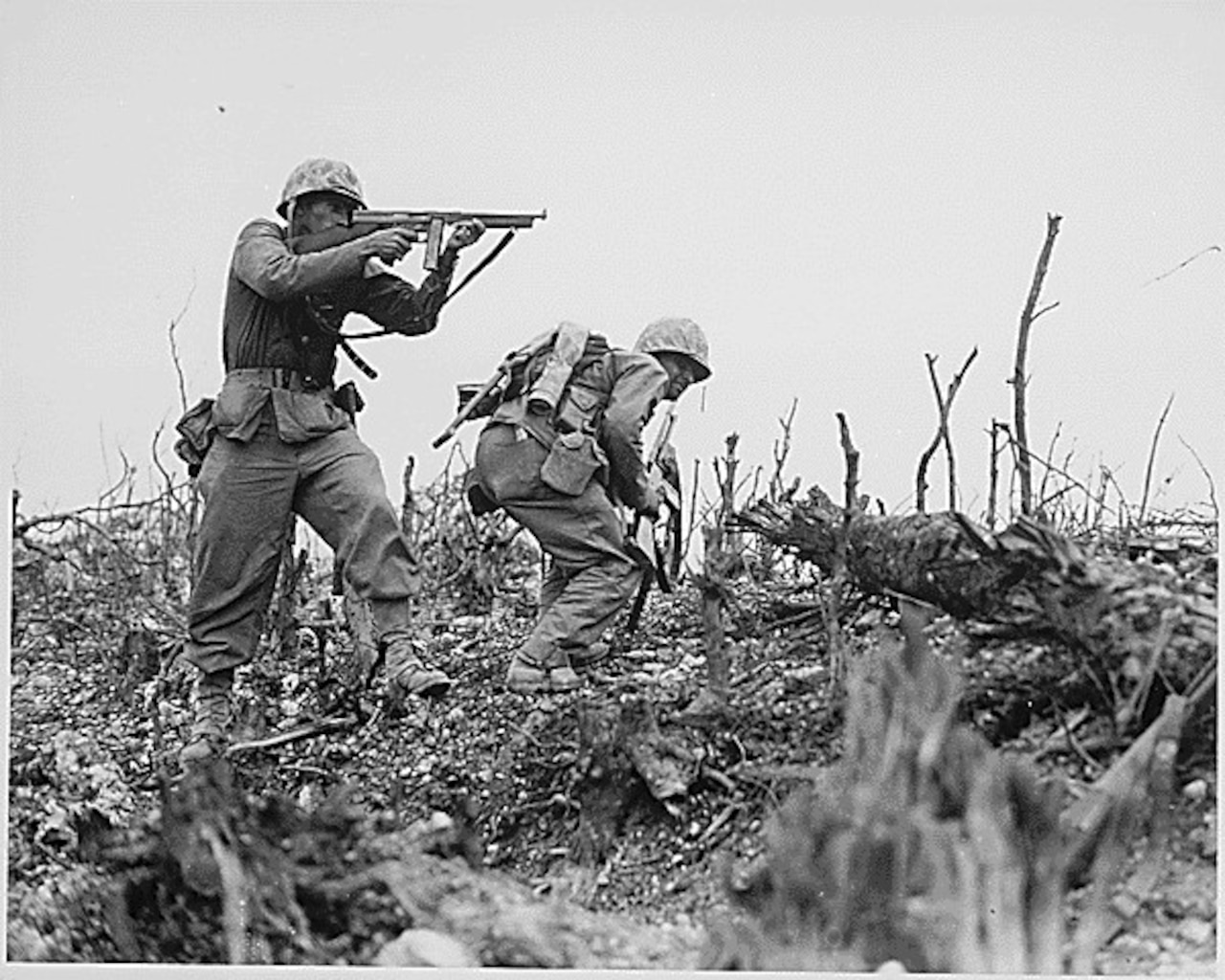 One man points his rifle as another man ducks down in brush.