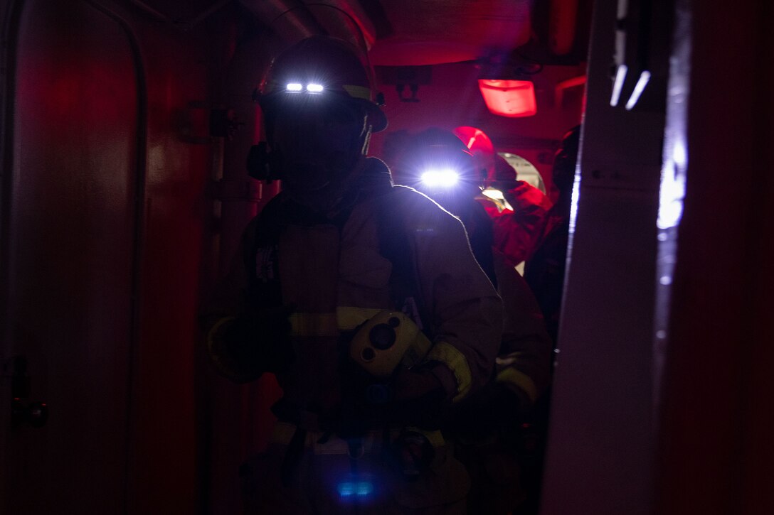 Sailors line up in firefighting gear in a dimly lit area of a ship.