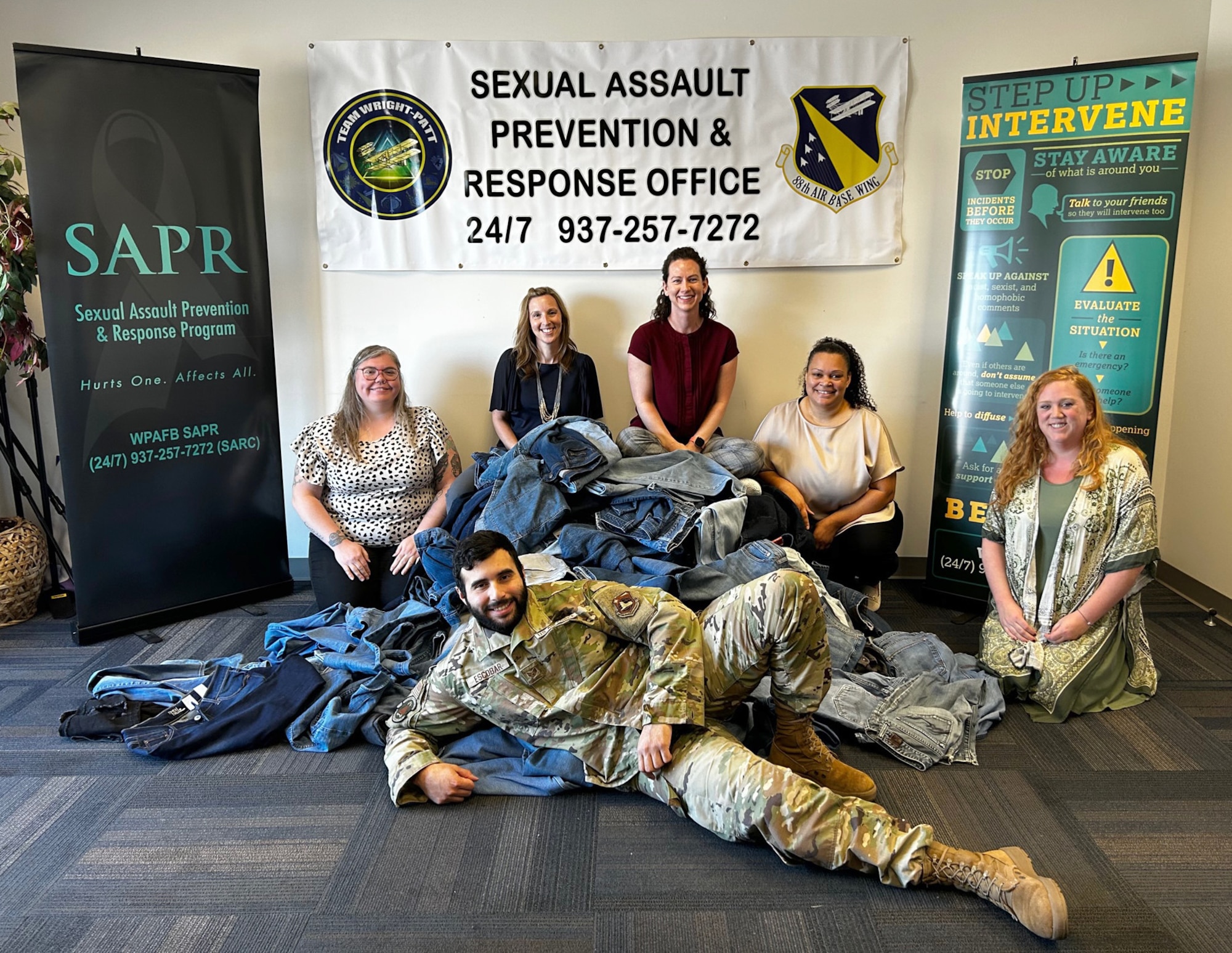 SAPR teammates pose next to a pile of denim with a sign in the background that reads Sexual Assault Prevention & Response Office.