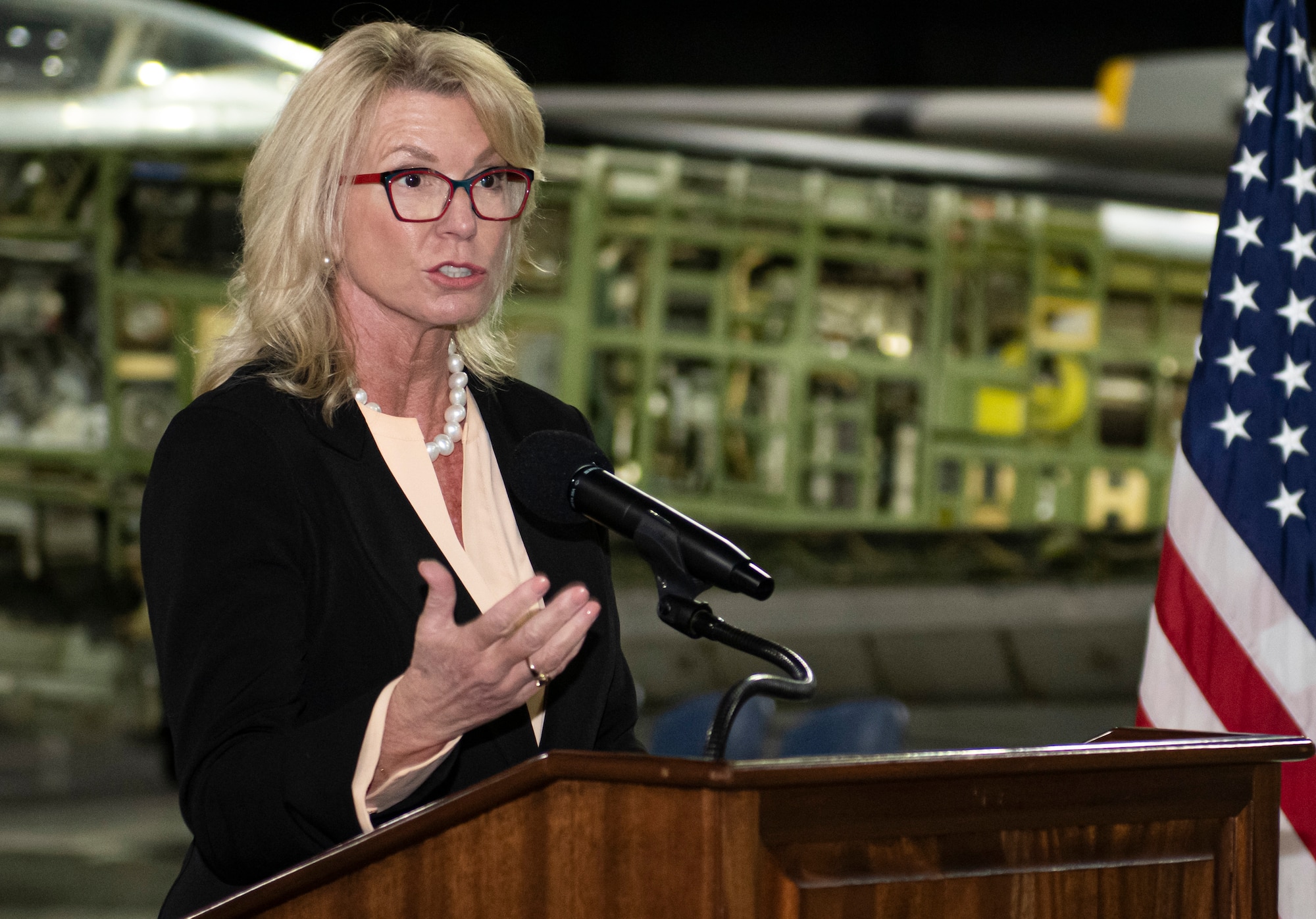 Woman speaking into a microphone at a podium, with the U.S. flag to the right.