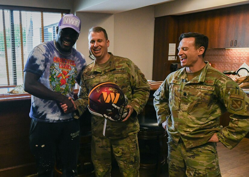 A man in civilian clothes gives a football helmet to two men in military uniforms.