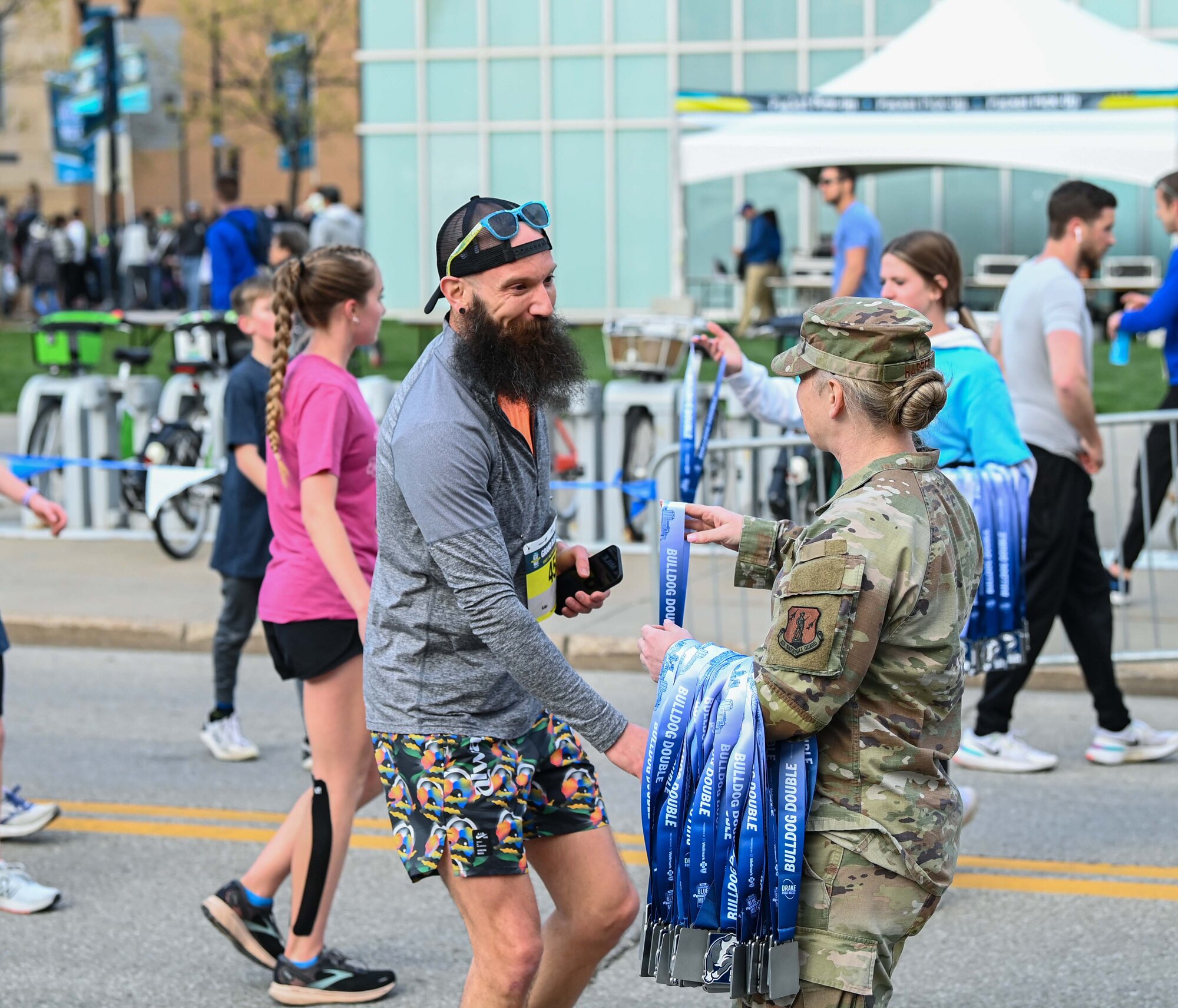 Female in green uniform hands blue medal to man with black beard wearing running clothes.
