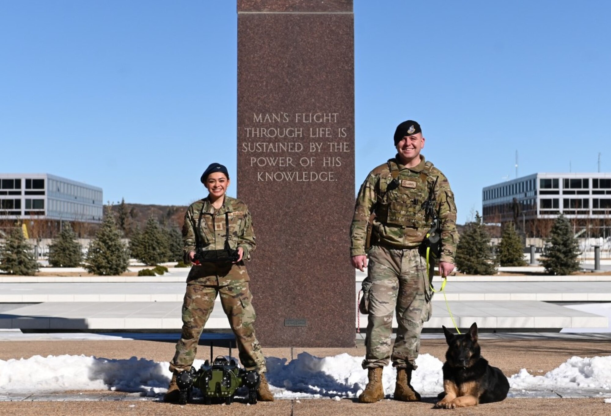 A female Cadet and male Security Forces Military Working Dog handler pose in front of the U.S. Air Force Academy's Eagle and Fledging Statue with a robot and Military Working Dog, February 2nd 2023.