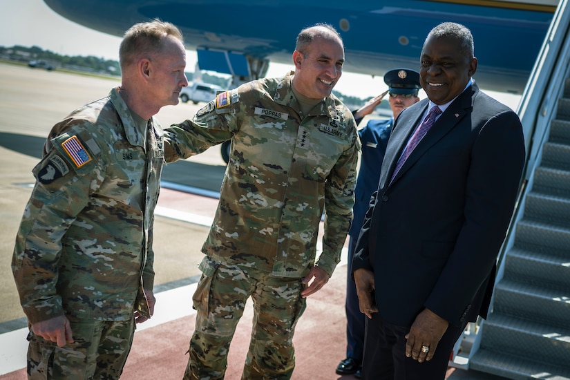 Three men chat in front of an airplane. Two of the men are in military uniform.