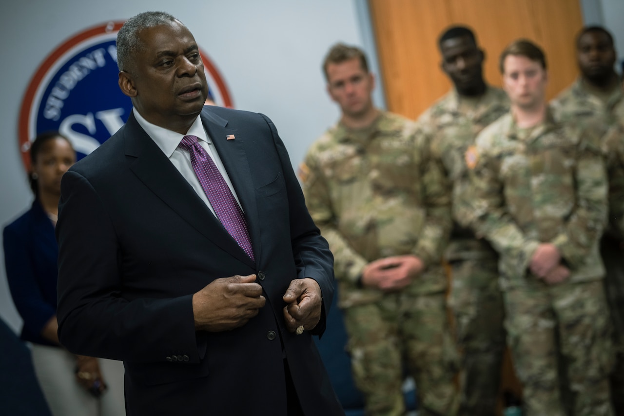 A man speaks while men in military uniform stand behind him.