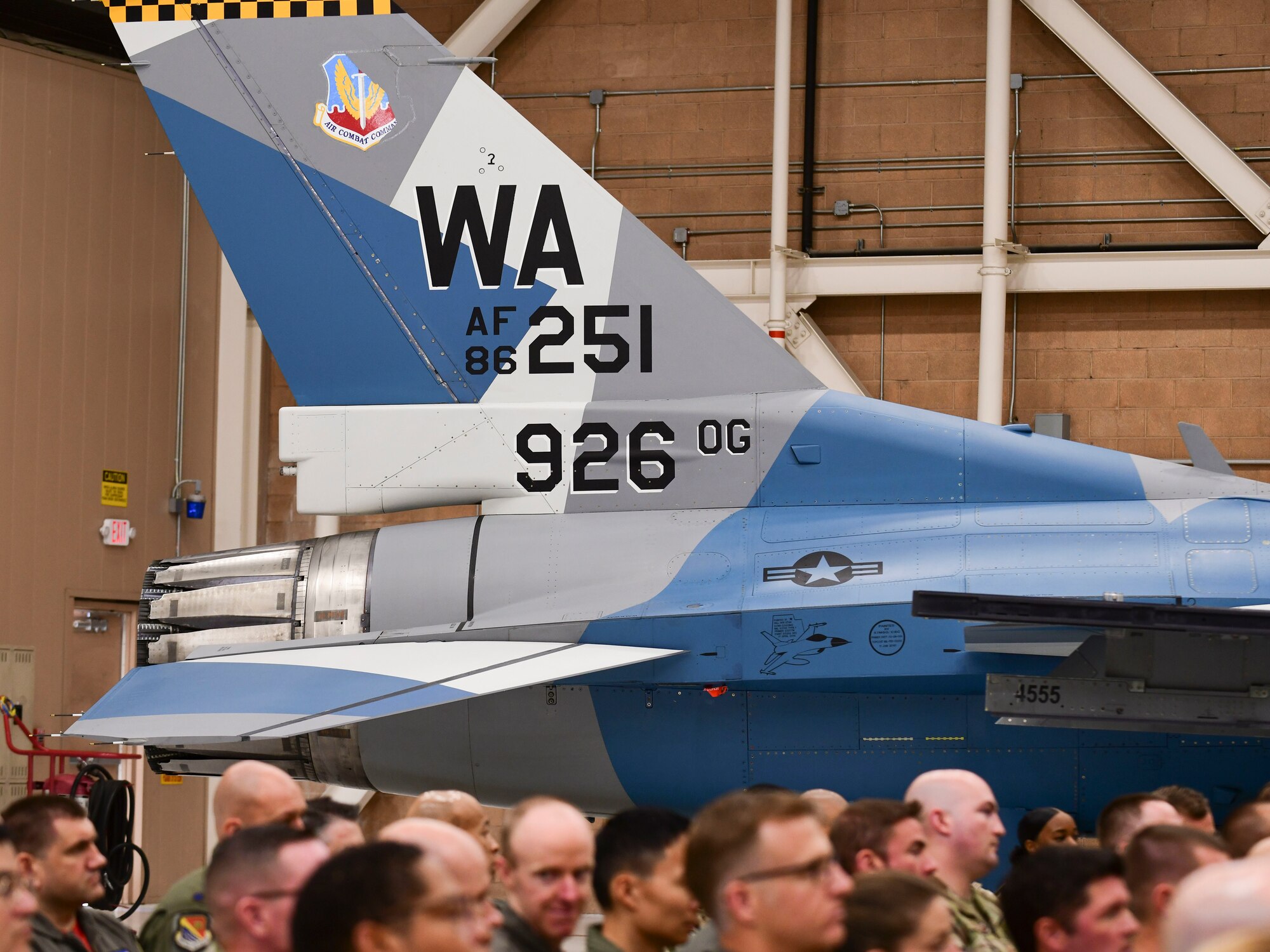 The back of a blue, gray and white-painted F-16 sits in a hangar behind the crowd during a ceremony. The tail of the aircraft reads in bold, black letters "WA, AF86, 251, 926 OG."