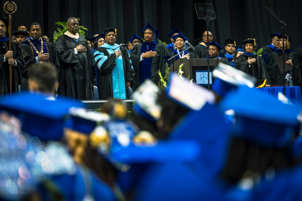 A group of people in gowns stand on a stage with hands over their hearts.