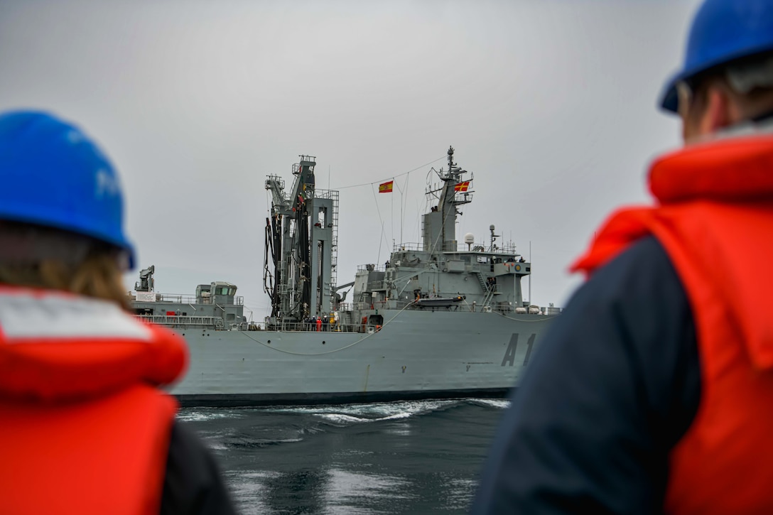 Two sailors watch as a ship sails close to their ship.