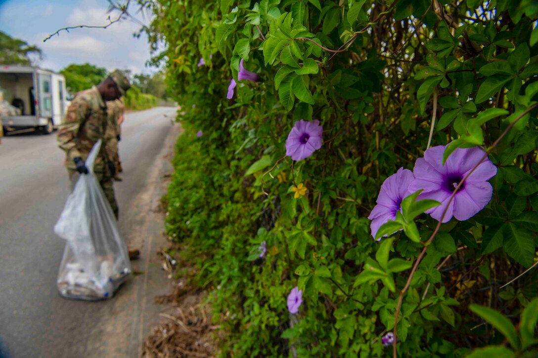 Airmen pick up trash next to a fence covered in vines and flowers.