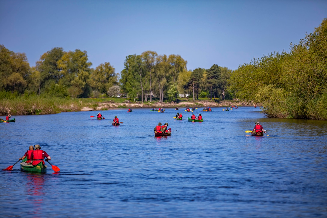 NATO soldiers wearing red life vests paddle canoes down river surrounded by greenery.