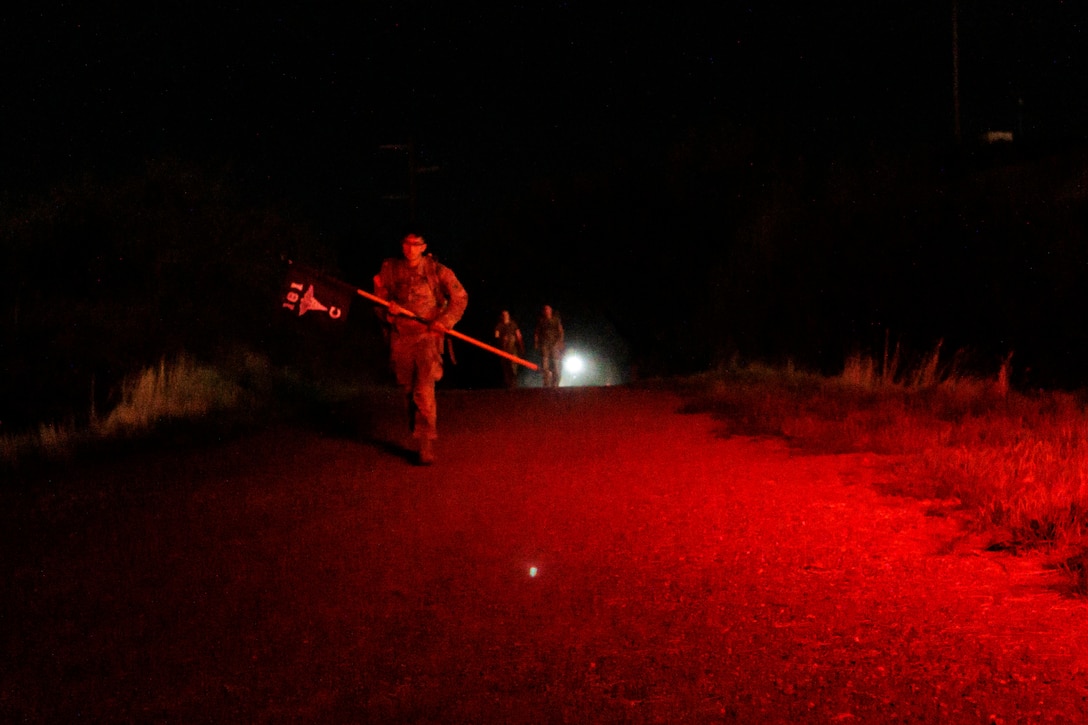 National Guardsmen carrying a rucksack march in a field in the dark illuminated by red light.