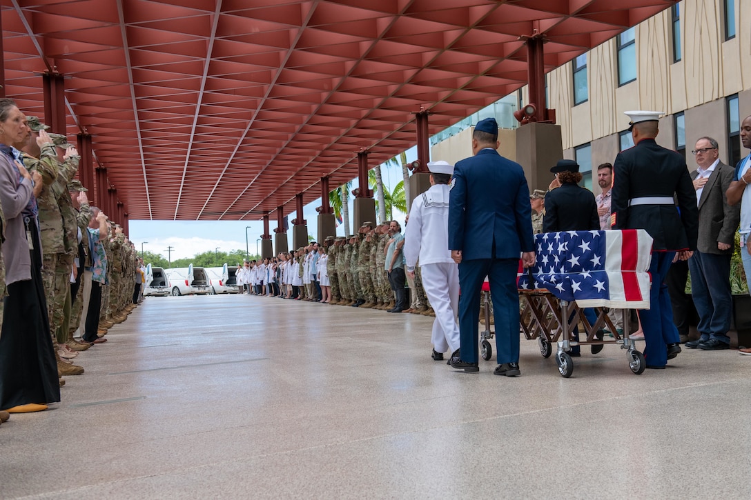 Service members walk beside a flag-covered casket as two lines of people salute or cover their hearts.