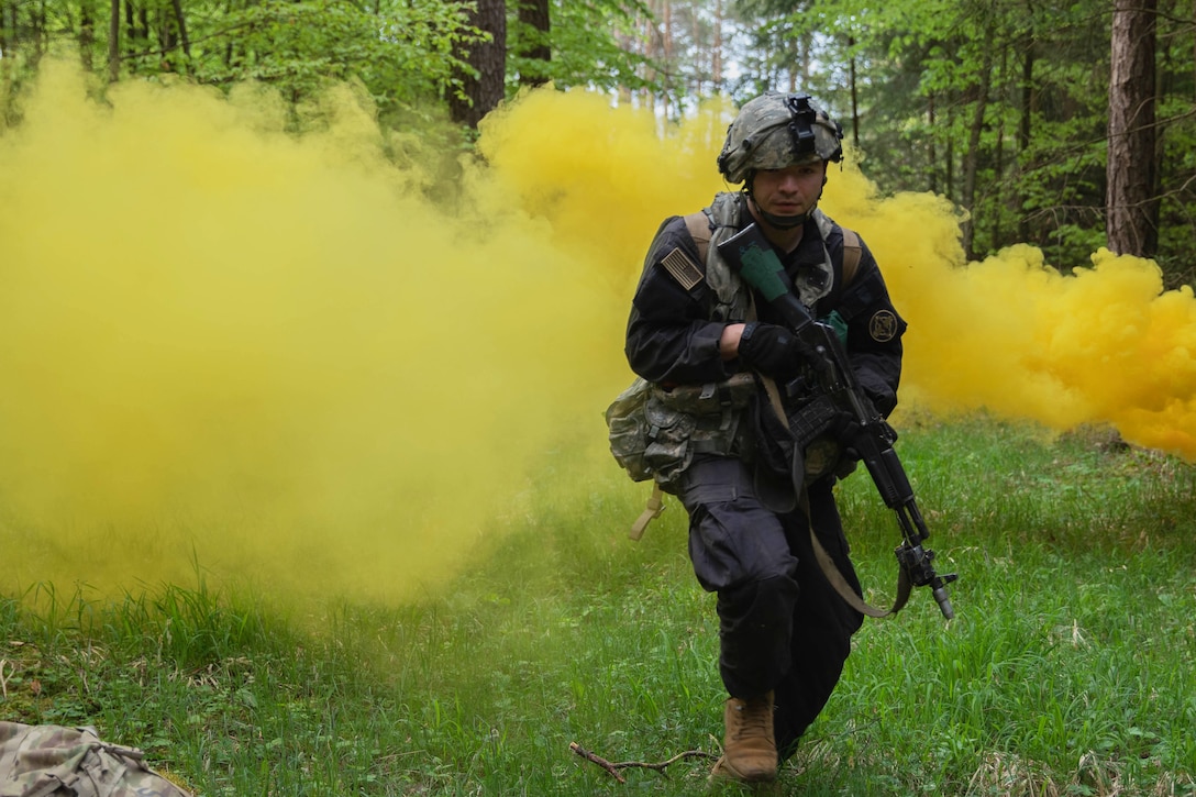 A soldier carrying a weapon and in full military gear runs through a field with green smoke in the background.