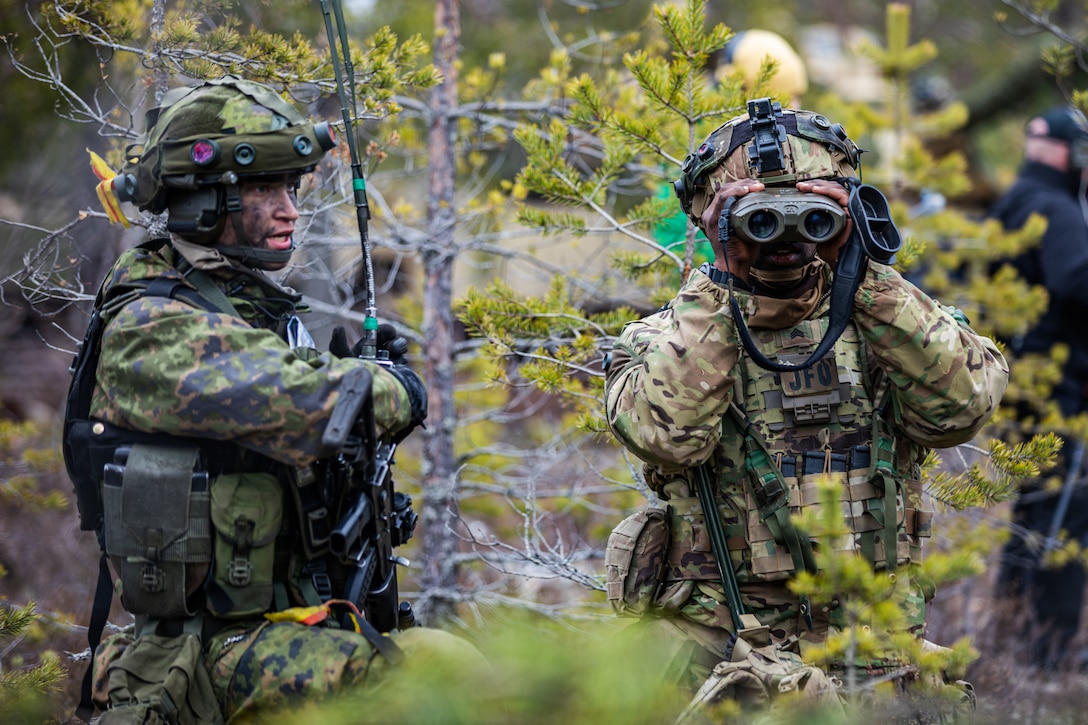 One soldiers peers through binoculars while another soldier looks on.