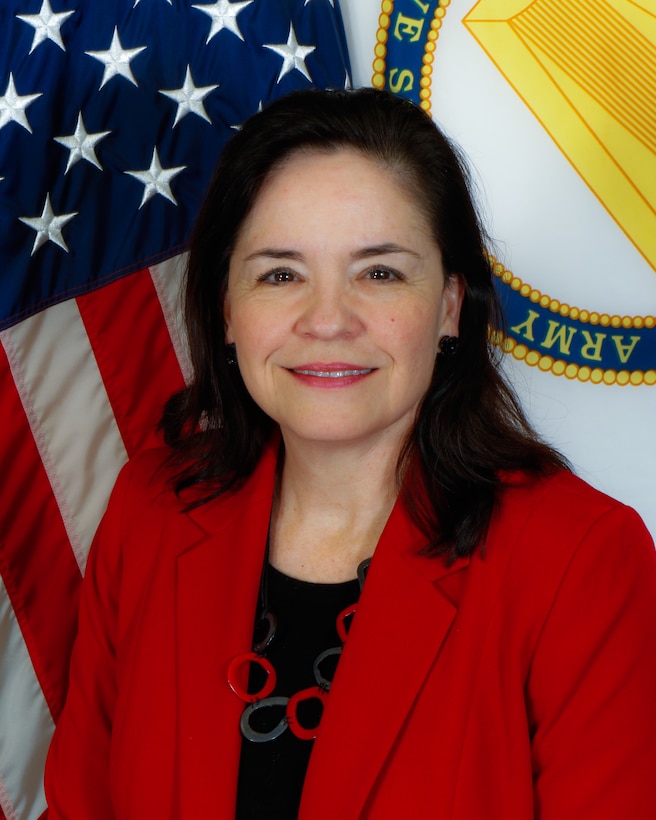 A woman, Ms. Karen Pane, in a red jacket and black blouse, sit in front front of the U.S. Flag and a white, yellow and blue flag representing the U.S. Army Corps of Engineers.